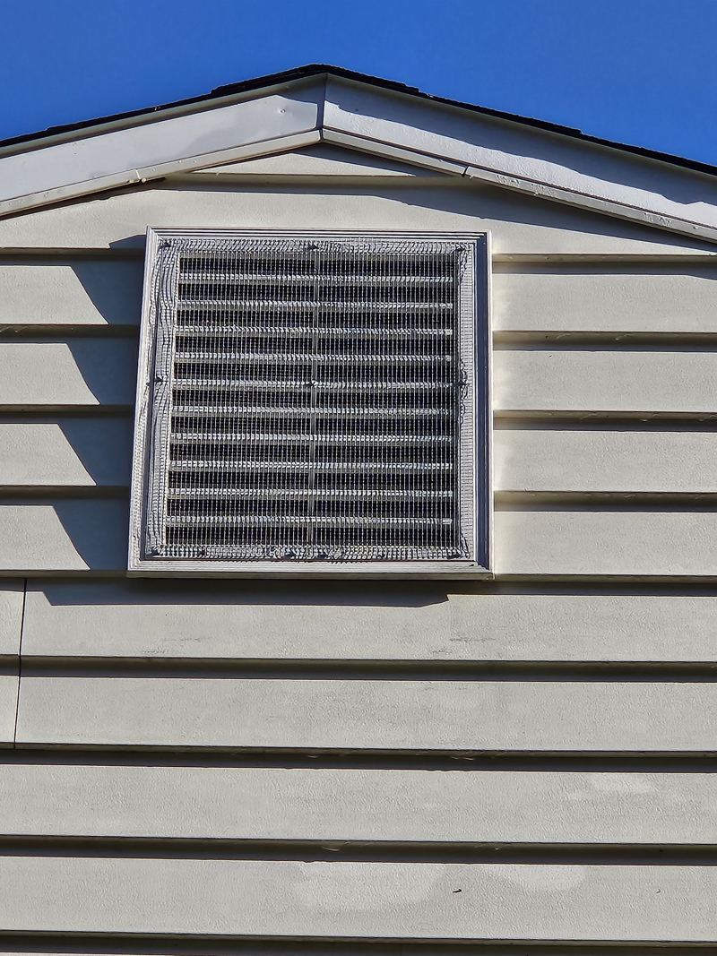 Mesh screening fitted over a gable vent on cream siding against blue sky